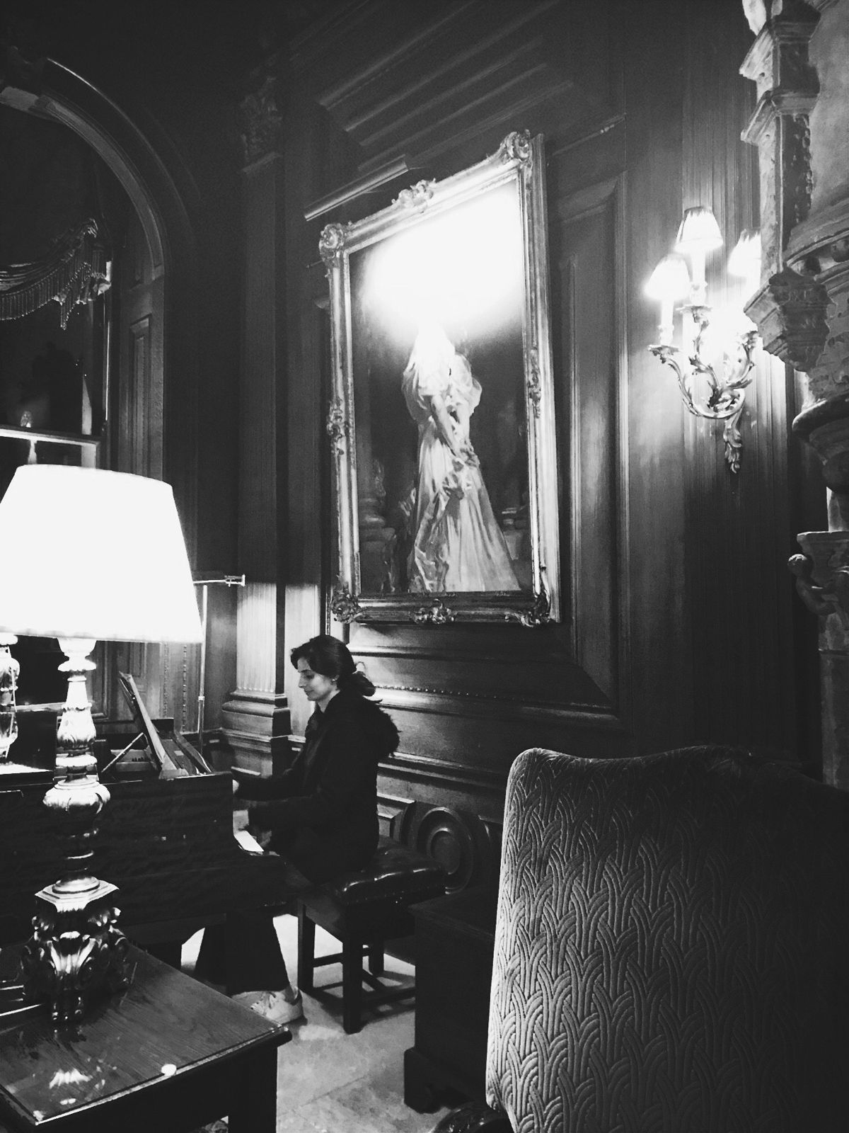 Naina at the piano beneath a large gilt-framed portrait, lamplit drawing room